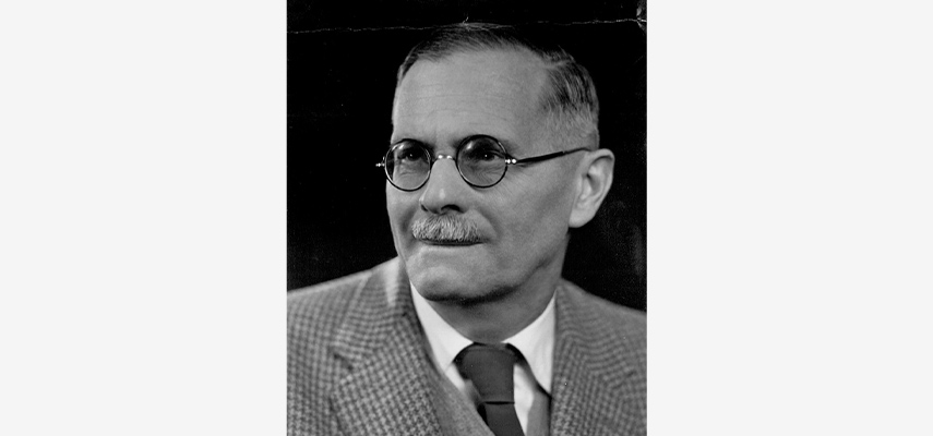 Schwarz-weißes Porträtfoto  von ein Mann mit Schnauzer, Brille, Anzug und Krawatte.