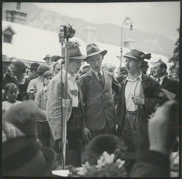 Schwarz-weißes Foto von Menschenmenge, vorne stehen drei Sänger mit Hut und Tracht.
