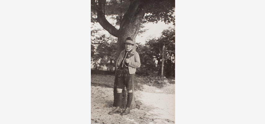 Schwarz-weißes Foto von Mann in Tracht, der an einem Baum lehnt.
