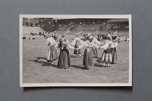 Eine Gruppe Tänzerinnen in Tracht steht im Kreis, dahinter sind Tribünen zu sehen.