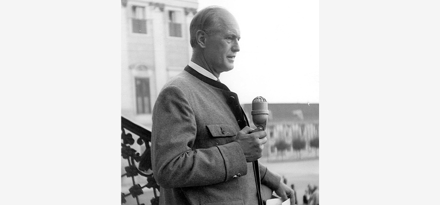 Schwarz-weißes Foto von einem Mann mit Halbglatze in einem Trachten-Anzug, der von einer Treppe aus in ein altmodisches Mikrofon spricht.