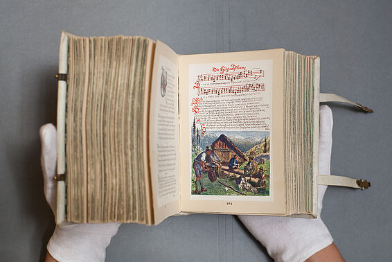 Aufgeschlagenes, kleines, dickes Buch. Die rechte Seite zeigt Musiknoten und Liedtext, darunter eine Zeichnung von einigen Männern vor einer Berghütte.