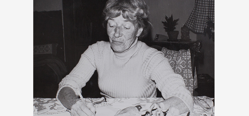 Frau mit kurzen Haaren und hellem Rollkragenpullover sitzt an einem Tisch und schreibt etwas auf.