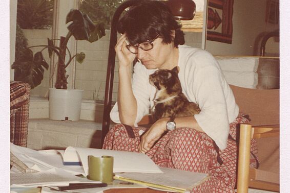 Älteres Foto von Frau mit schwarzen Haaren und Brille, die mit Katze auf dem Schoß an einem Couchtisch sitzt und nachdenkt