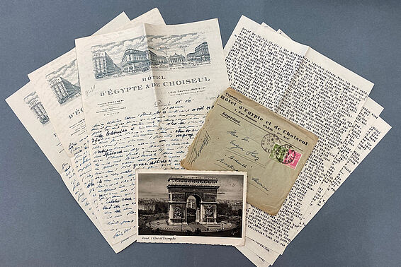 Verschiedene getippte und handgeschriebene Briefe, ein Foto vom Arc de Triomphe und ein zerrissener Briefumschlag liegen auf einer Tischplatte arrangiert