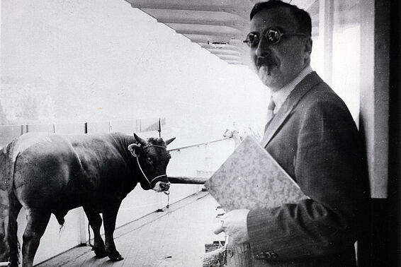 Schwarz-weißes Foto von Mann mit Brille und einer Mappe unter dem Arm neben einer Kuh