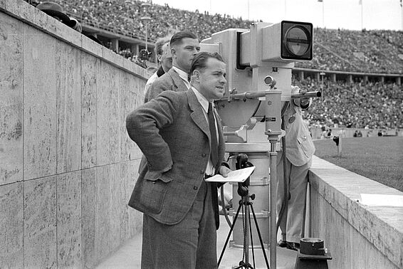 Vier Männer in Anzügen neben Filmkamera in einem Stadion