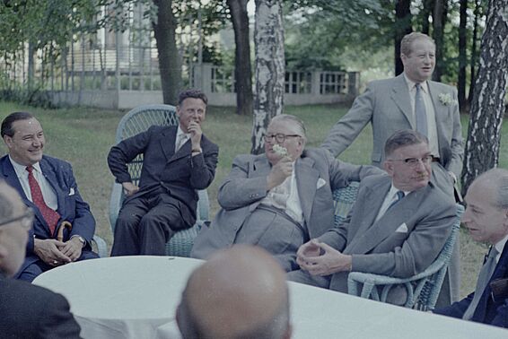 Gruppe von Männern in Anzügen sitzt um einen Gartentisch und lacht