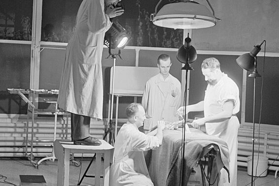 Drei Männer operieren an jemandem, ein Mann sieht von einem Stockerl aus zu.
