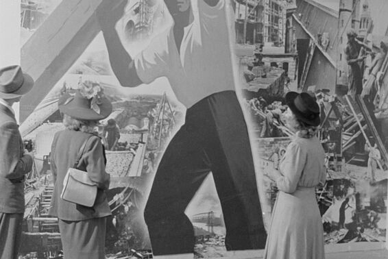 Zwei Frauen und ein Mann stehen vor einem Plakat, auf dem ein Arbeiter einen Balken hebt, im Hintergrund eine Stadt in Trümmern