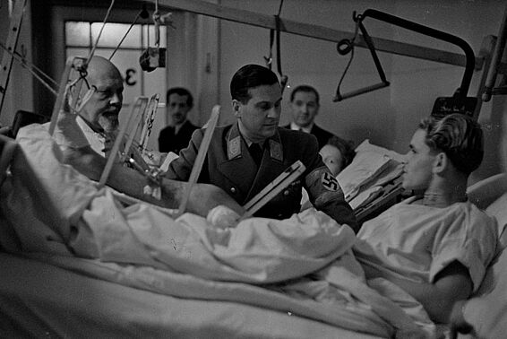 Ein Mann liegt in einem Krankenbett und redet mit zwei Männern, die neben dem Bett stehen