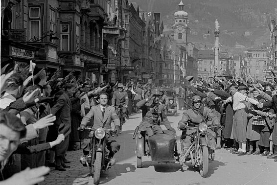 Drei Männer fahren auf Motorrädern durch ein Dorf, rechts und links stehen Menschenmengen, die den Hitlergruß machen, im Hintergrund Berge