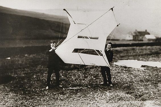 Zwei Männer in Anzügen stehen auf einer Wiese und halten einen sehr großen Segeldrachen in Händen, Schwarzweiß-Fotografie