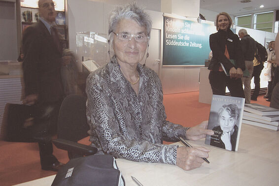Ruth Klüger mit kurzen grauen Haaren, Lesebrille und Bluse mit schwarzweißem Schlangenprint, und Buch in der Hand auf einer Lesung