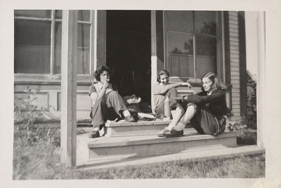 Schwarz-weißes Foto von drei jungen Menschen, die auf einer Veranda sitzen. Eine von ihnen raucht eine Zigarette.
