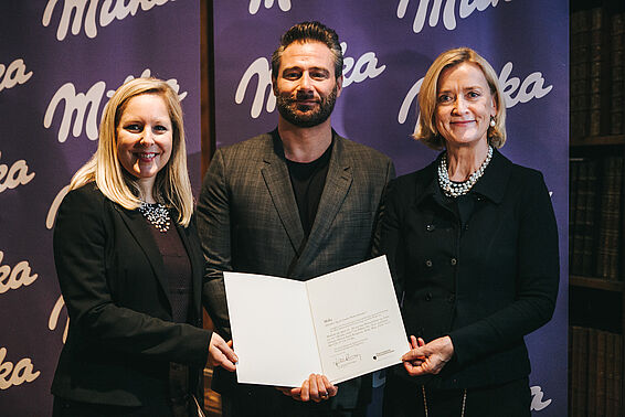 Zwei Frauen und ein Mann in eleganter Kleidung halten eine Urkunde vor einer Wand mit dem Milka-Logo