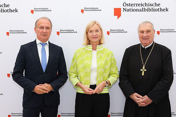 Ein Mann im Anzug, eine Frau im gelben Sakko und ein Priester mit Kreuz um den Hals