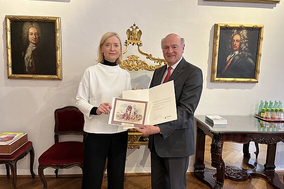 Eine blonde Frau mit weißem Oberteil und ein glatzköpfiger Mann im Anzug halten eine Urkunde in die Kamera
