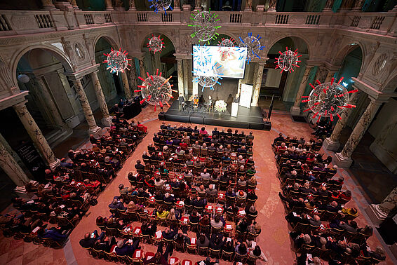 Volles Publikum vor Bühne in prunkvollem Saal.