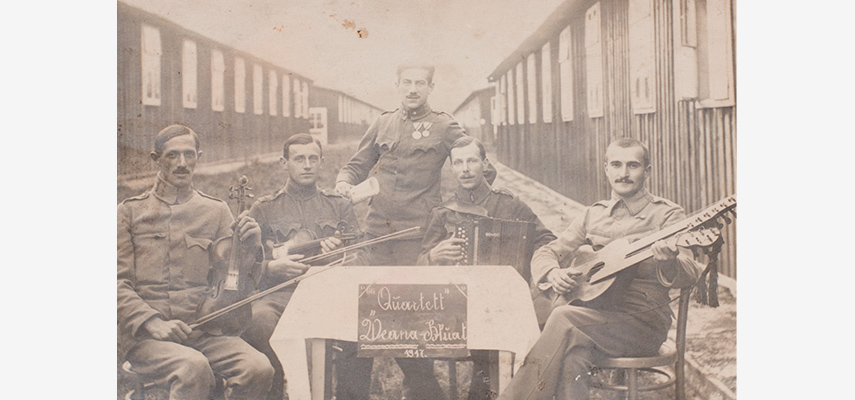 Schwarz-weißes Foto von fünf Männern. Vier davon halten Musikinstrumente und sitzen an einem Tisch, in der Mitte hinter dem Tisch steht der fünfte. Alle tragen Uniform.