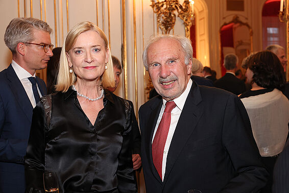 Eine Frau und eine Mann in eleganter Kleidung posieren stehend, im Hintergrund Gäste