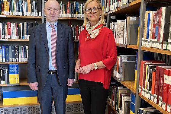 Ein Mann im Anzug und eine Frau mit rotem Oberteil stehen vor Bücherregalen, die mit der ukrainischen Flagge dekoriert wurden.