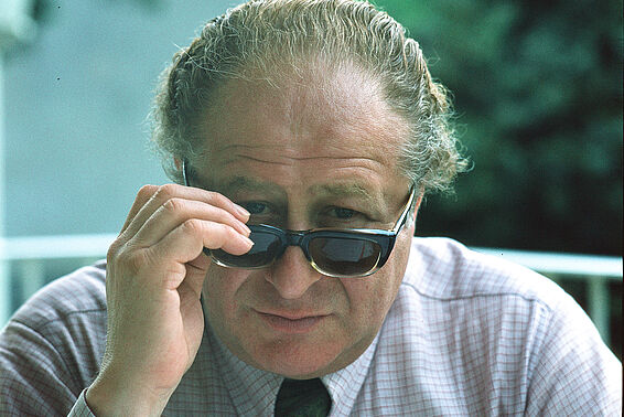 Foto von Mann mit zurückgekämmten Haaren in Hemd und Krawatte, der gerade seine Sonnenbrille abnimmt