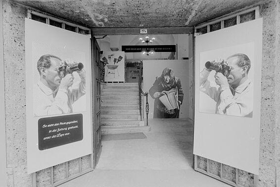 Bilder eine Fotoausstellung, Mann schaut durch Fotokamera, eine Frau mit Kopftuch hält einen Stapel Zeitungen in der Hand