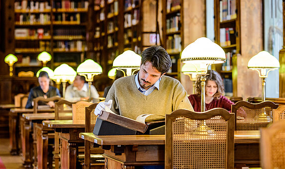 Fotografie des Augustinerlesesaal der Österreichischen Nationalbibliothek der Österreichischen Nationalbibliothek, im Zentrum ist ein Mann zu sehen, der in einer historischen Handschrift liest.