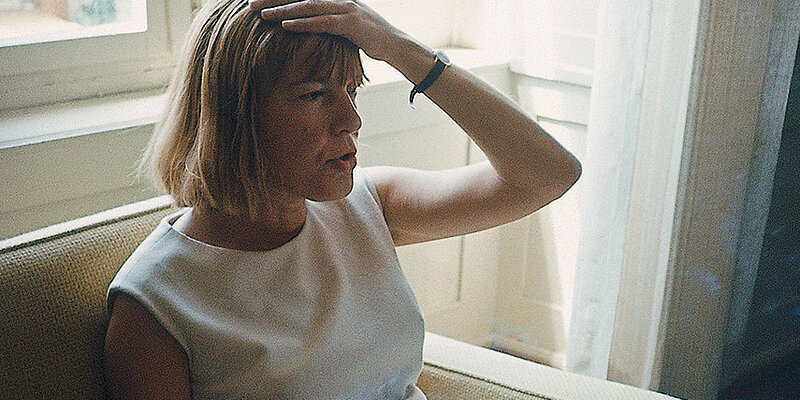 Foto von Ingeborg Bachmann, die auf einem Sofa sitzt und sich an den Kopf greift. 