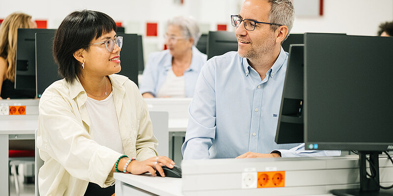 Eine jüngere Person und eine älterer Person sitzen an einem Computer.