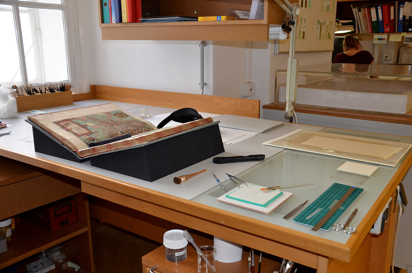 Auf einem Arbeitsplatz liegt eine geöffnete kolorierte Druckschrift auf Keilkissen. Alle Materialien und Werkzeuge liegen bereit, um durch Kupferfraß brüchig gewordene Stellen zu hinterkleben.