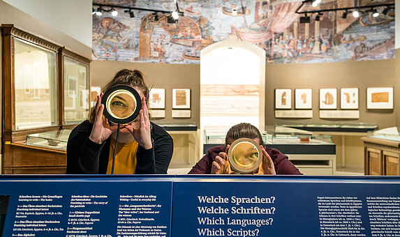 Zwei Personen stehen im Papyrusmuseum und blicken durch eine Lupe direkt in die Kamera.