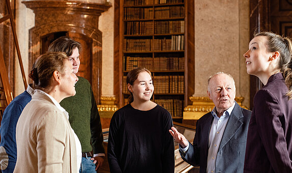 Eine Gruppe von Menschen nimmt an einer Führung in einem barocken Bibliothekssaal teil.