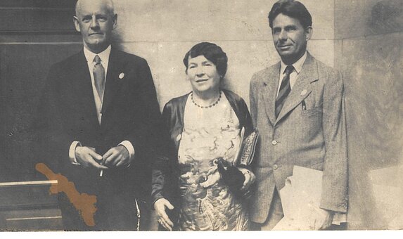Schwarz-weißes Foto von drei Personen in eleganter Kleidung vor einem Gebäude.