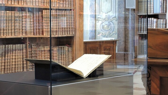 Eine Vitrine im Prunksaal in dem eine aufgeschlagene Musikhandschrift liegt. Im Hintergrund sind Buchregale des Prunksaals zu sehen.