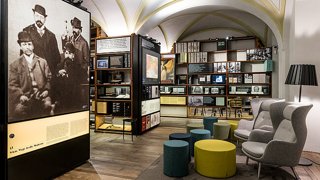 Ein Blick in das Literaturmuseum. Die Regalstruktur ist gefüllt mit Objekten und großflächigen Abbildungen, gemütliche Sitzgelegenheiten laden zum Verweilen ein. 