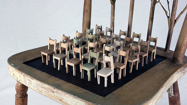 Das Bild zeigt einen Sessel, auf dem dutzende sehr kleine Sessel platziert sind. 