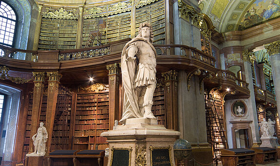 Foto von Statue in barockem Saal mit hohen Bücherregalen und Fresken.