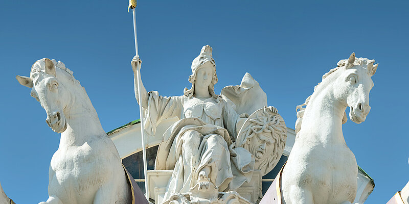 Foto von weiblicher Statue in griechischer Kleidung, Speer, Schild, mit Pferden rechts und links.
