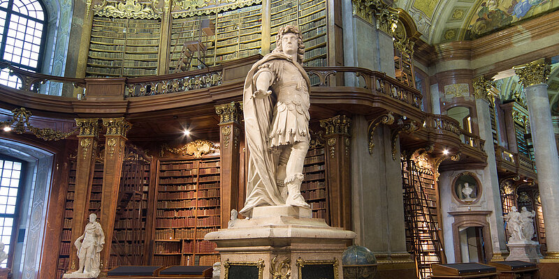 Foto von Statue in barockem Saal mit hohen Bücherregalen und Fresken.