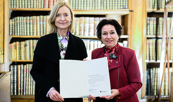 Zwei Personen nebeneinander halten zusammen eine Urkunde, im Hintergrund ein Bücherregal.