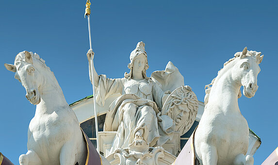 Foto von weiblicher Statue in griechischer Kleidung, Speer, Schild, mit Pferden rechts und links.