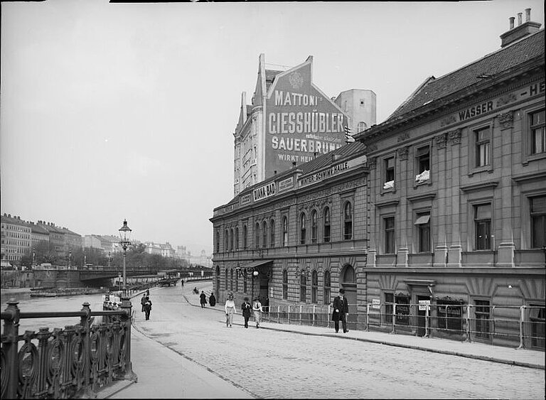 Schwarz-weißes Foto von Brücke in einer Stadt mit kleineren Gebäuden und ein paar Personen. Auf einem Gebäude steht "Dianabad", an einem höheren Gebäude dahinter ist eine Reklame.