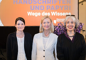 Drei Personen in eleganter Kleidung vor einer Leinwand. 