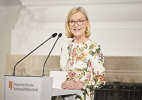 Person im eleganten Blazer an Rednerpult.