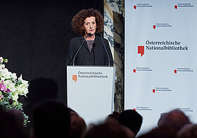 Person am Rednerpult blickt zum Publikum.  