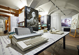 Museumsraum mit Regalen mit Fotografien, im Vordergrund vier Schreibmaschinen.