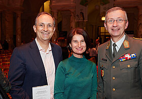 Zwei Personen in eleganter Kleidung und Person in Militär-Uniform posieren lächelnd. 