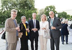 Fünf Personen in eleganter Kleidung mit Weingläsern in der Hand stehen im Freien. 